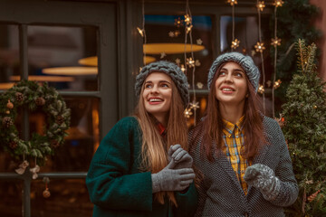 Christmas, New Year, winter holidays conception: two happy smiling women, friends, sisters, wearing knitted hat, coats, gloves, posing in street with festive decorations. Copy, empty space for text