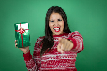 Young beautiful girl holding gift over isolated white background laughing at you, pointing finger to the camera with hand over body, shame expression