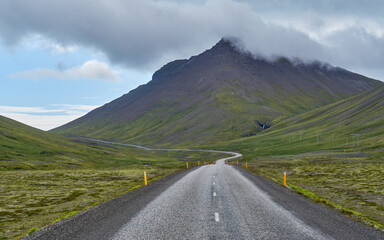 A scenic road stretches toward a cloud-topped cone-shaped mountain between Hofn and Djupivogur in...
