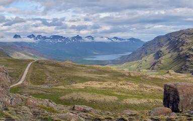 A winding gravel backroad snakes through rolling hills and valleys in rural Iceland, flanked by...