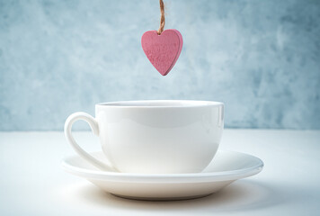 A pink heart hangs over a coffee Cup and saucer on a white and gray background. Side view. Concept February 14.