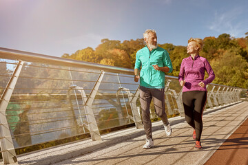 Stay in shape. Full length shot of active mature family couple in sportswear smiling at each other while jogging together on a sunny autumn day. Joyful senior couple doing sport outdoors