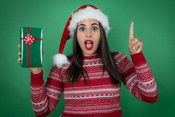 Young beautiful girl wearing Christmas Santa hat holding gift over isolated white background amazed...