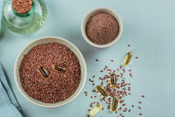 Brown flax seed or linseed and ground or crushed flaxseed in small bowl and gelatin capsules with omega oil on a light blue background.