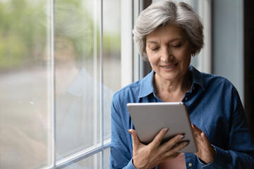 Close up smiling mature woman using tablet, standing near window at home, satisfied gray haired female looking at device screen, shopping or chatting with relatives online, browsing apps