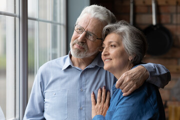 Happy dreamy mature couple hugging, thinking about good future together, standing in kitchen, looking out window, smiling senior husband wearing glasses embracing wife enjoying tender moment