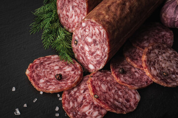 Sliced salami sausage on a board. Meat product and herbs on a dark back. A quick meal or snack. Dried sausage with black peppercorns.