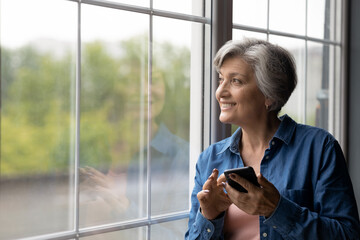 Smiling dreamy mature woman holding phone, looking out window close up, distracted from chatting...