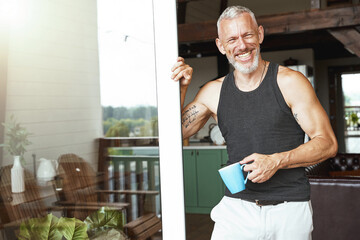 Perfect morning. Handsome positive middle aged caucasian man with coffee standing near terrace entrance of his modern apartment