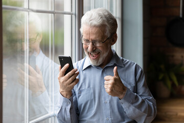 Close up overjoyed mature man using phone, reading good news in message, showing thumb up, standing...