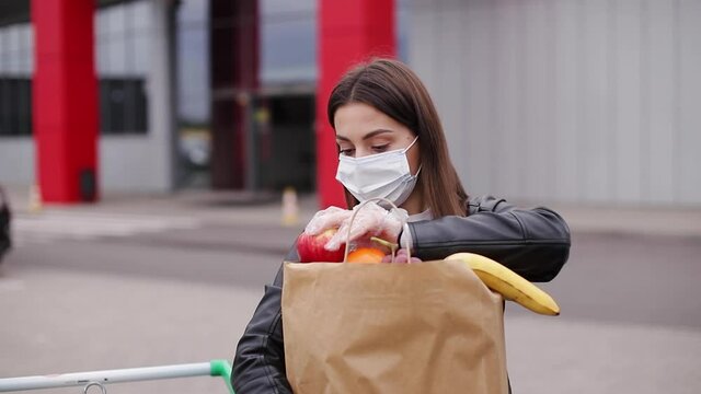 Groceries in female's hand by the car truck. Woman in protective mask and disposable gloves. Fresh fruits in eco paket. Food shopping during coronavirus Covid-19 quarantine. Woman take apple to feel