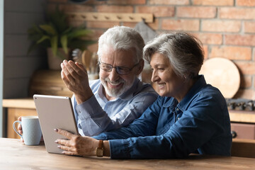 Happy mature couple having fun with tablet, sitting in kitchen, laughing senior man wearing glasses drinking tea or coffee in morning, grandparents chatting with relatives online, browsing apps