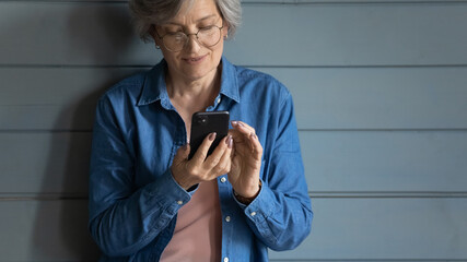Close up isolated mature woman wearing glasses using smartphone, standing on grey wooden wall background, shopping or chatting with relatives online, browsing mobile device apps, generation and tech