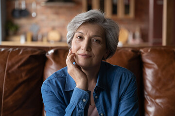 Head shot portrait mature woman sitting on couch at home alone, attractive focused senior middle aged grey haired female looking at camera, posing for photo, relaxing on cozy sofa indoor