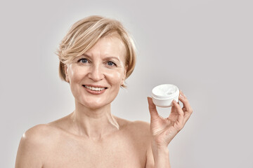 Portrait of beautiful middle aged woman smiling at camera, holding moisturizing facial cream while posing isolated over grey background