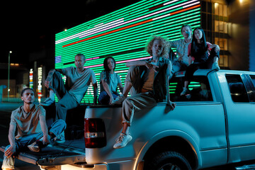 Fototapeta na wymiar Six casually dressed youngsters of different nationalities sitting in an opened pick-ups trunk next to each other looking into a camera and posing with a huge led screen behind