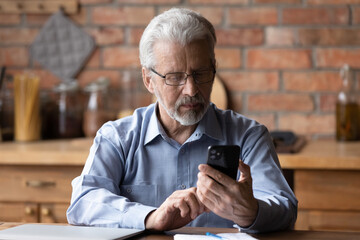 Close up focused senior man wearing glasses using phone, sitting at table in kitchen, serious...