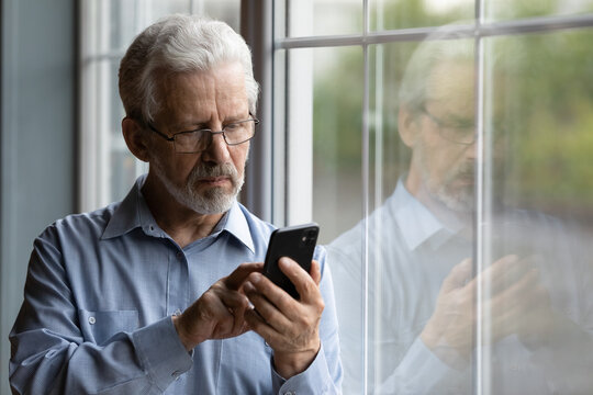 Close Up Serious Mature Man Wearing Glasses Using Phone, Standing Near Window At Home, Lonely Grandfather Waiting For Call From Relatives Or Grandchildren, Looking At Smartphone Screen