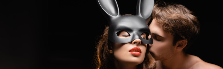 Shirtless man kissing sexy woman in rabbit mask with red lips isolated on black, banner