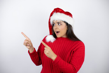 Young beautiful woman wearing a Santa hat over white background surprised and pointing her fingers side