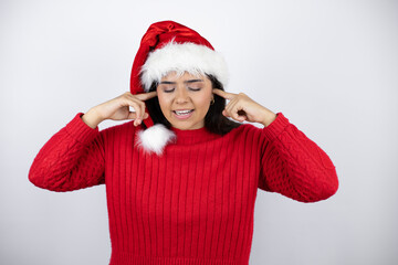 Young beautiful woman wearing a Santa hat over white background covering ears with fingers with...