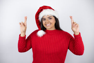 Young beautiful woman wearing a Santa hat over white background gesturing finger crossed smiling with hope