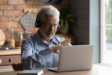 Close up mature man wearing headphones using laptop, making video call, sitting at table in...