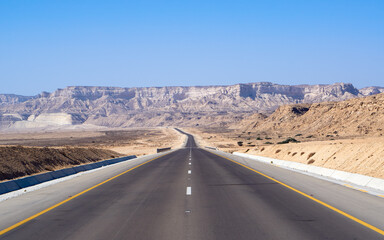 An asphalt highway cuts through the rugged Dhofar plateau in Oman, flanked by rocky cliffs and desert slopes under a clear blue sky.
