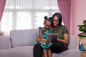 Mother was teaching the daughter through tablet joyfully while sitting on the sofa. black people or African Americans. Home study concept. Parent-teacher concept.