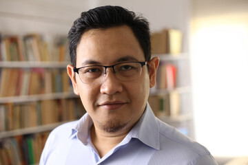 Portrait of cheerful Asian male librarian smiling, man standing against books in library