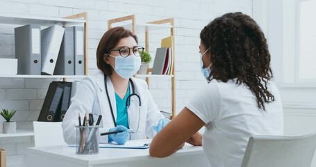Woman doctor in medical mask asking female patient about symptoms, writing personal medical history at clinic