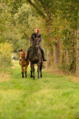 Fototapeta premium Young woman riding without saddle on her beautiful brown mare, yellow foal next to them, in the autumn forest