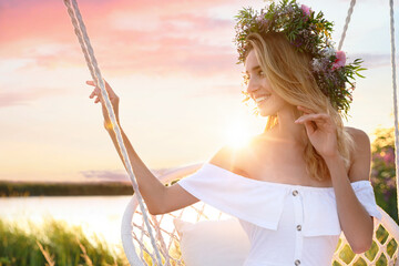 Young woman wearing wreath made of beautiful flowers on swing chair outdoors