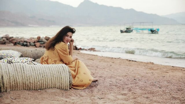 Attractive brunette woman sitting on a deserted beach at windy weather. Gorgeous slim mixed race Asian Caucasian girl in yellow dress spending holidays on the sea shore.