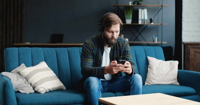 Young attractive Male is enjoying the Music, wearing Headphones, and texting someone through an App on his Phone. Portrait of a young man spending time, having Fun at Home. Apps. Music. Indoor.
