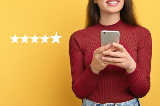 Woman leaving review online via smartphone on yellow background with five stars, closeup. Service or product feedback