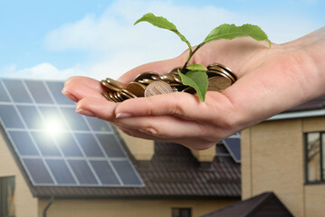 Woman holding coins and green sprout against house with installed solar panels on roof, closeup....