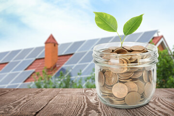 Glass jar with coins and plant against house with installed solar panels on roof. Economic benefits of renewable energy