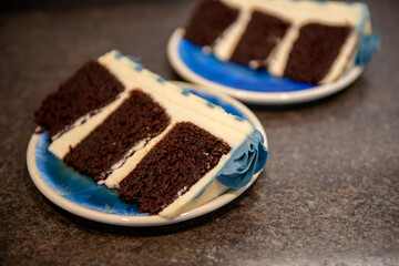 Two pieces of sliced chocolate cake with white icing and blue flowers. The sweet dessert is a three layer birthday cake. The blue dessert plates are laying on a textured countertop.