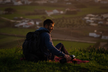 Naklejka na ściany i meble Hiker man sitting on the ground in mountains and pouring tea from thermos to cup