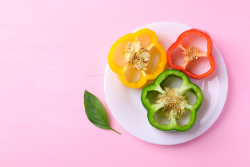 Sliced green, yellow and red bell peppers on white plate with leaf on pink background, Top view