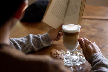 Man holding a coffee cappuccino cup while reading a book, leaning his arms on a wooden table with a black beret on it