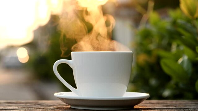 Close-up steaming hot coffee or tea cup on saucer, slow motion. Hot ceramic white coffee cup with smoke on old wooden table in nature background. Hot Coffee Drink Concept