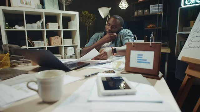 Portrait of african american guy working with papers near laptop in dark office.