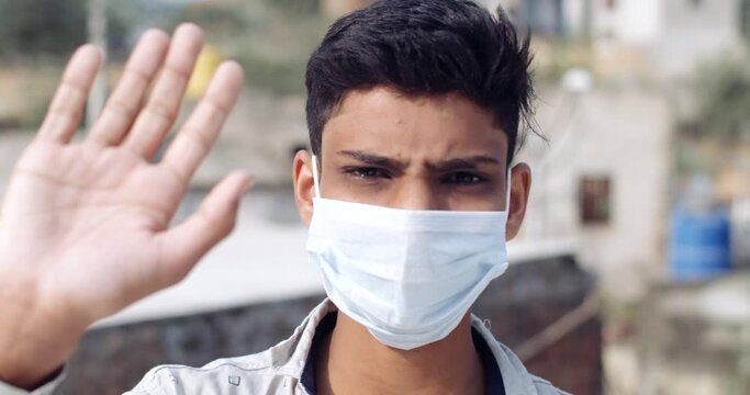 Slow-motion CU of an Indian teen boy wearing protective face mask against covid19 outdoors making hand gestures as he waves his hand goodbye staring at camera pov 