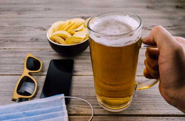 glass of beer, crisps, sunglasses, cell phone and face mask on a wooden table