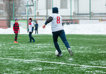 Boys in white sportswear running on soccer field with snow on background. Young footballers dribble and kick football ball in game. Training, active lifestyle, sport, children winter activity