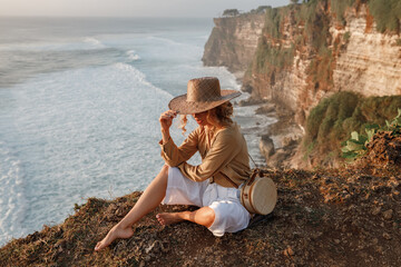 Young tourist  woman in straw hat, cover the face, sit  on the edge of a cliff with sea view. Travel Bali, explore beautiful places of the island