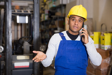 Foreman in protective helmet and overalls talking on cell phone in warehouse of a hardware store