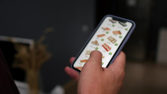A man at home, in the living room, buys sushi from an online store for a courier to deliver it. A man orders food at home in an online store using a smartphone. A man chooses dinner at a restaurant